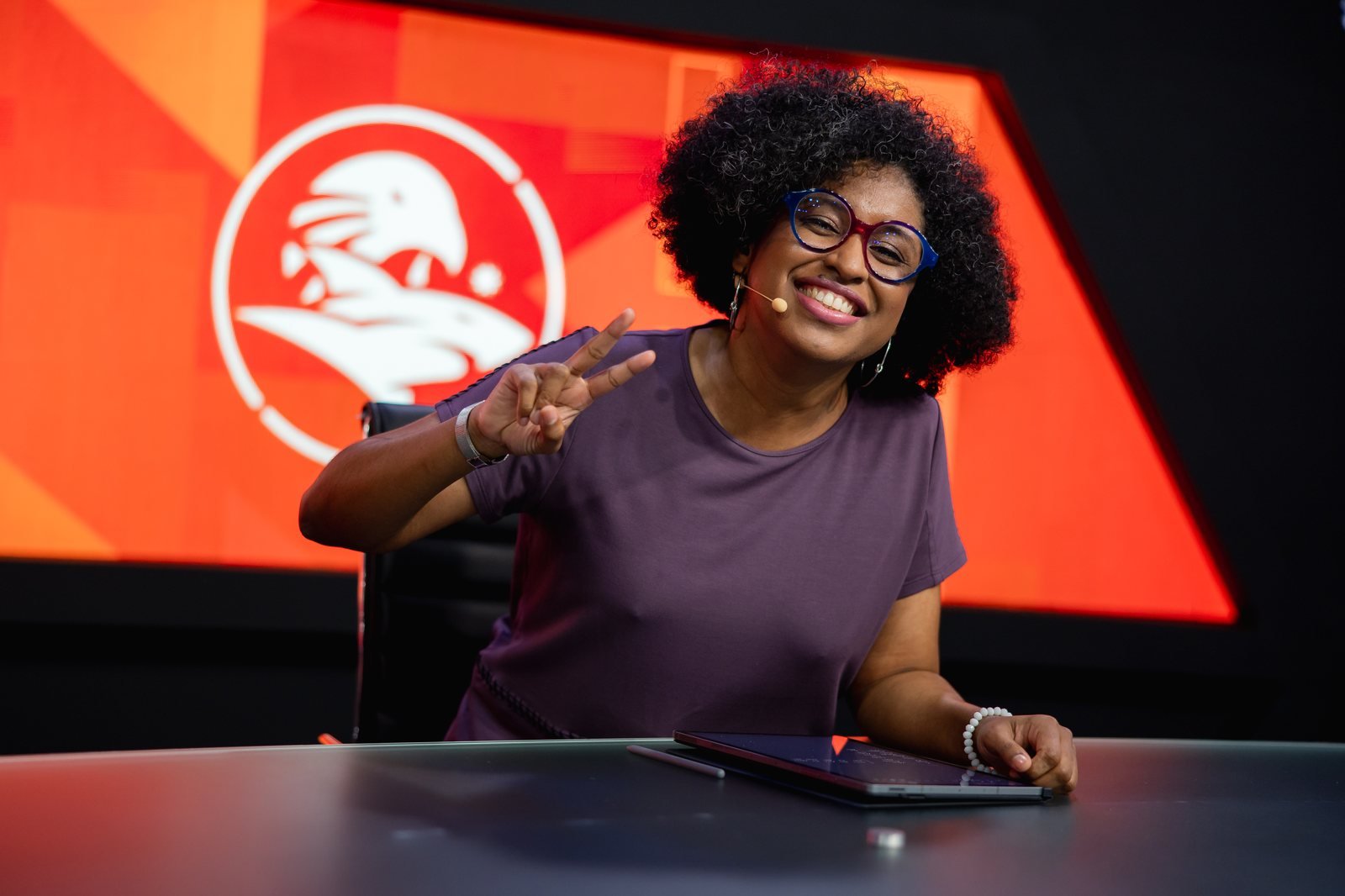 Lahgolas sorrindo em uma bancada de transmissão e fazendo o gesto de paz.